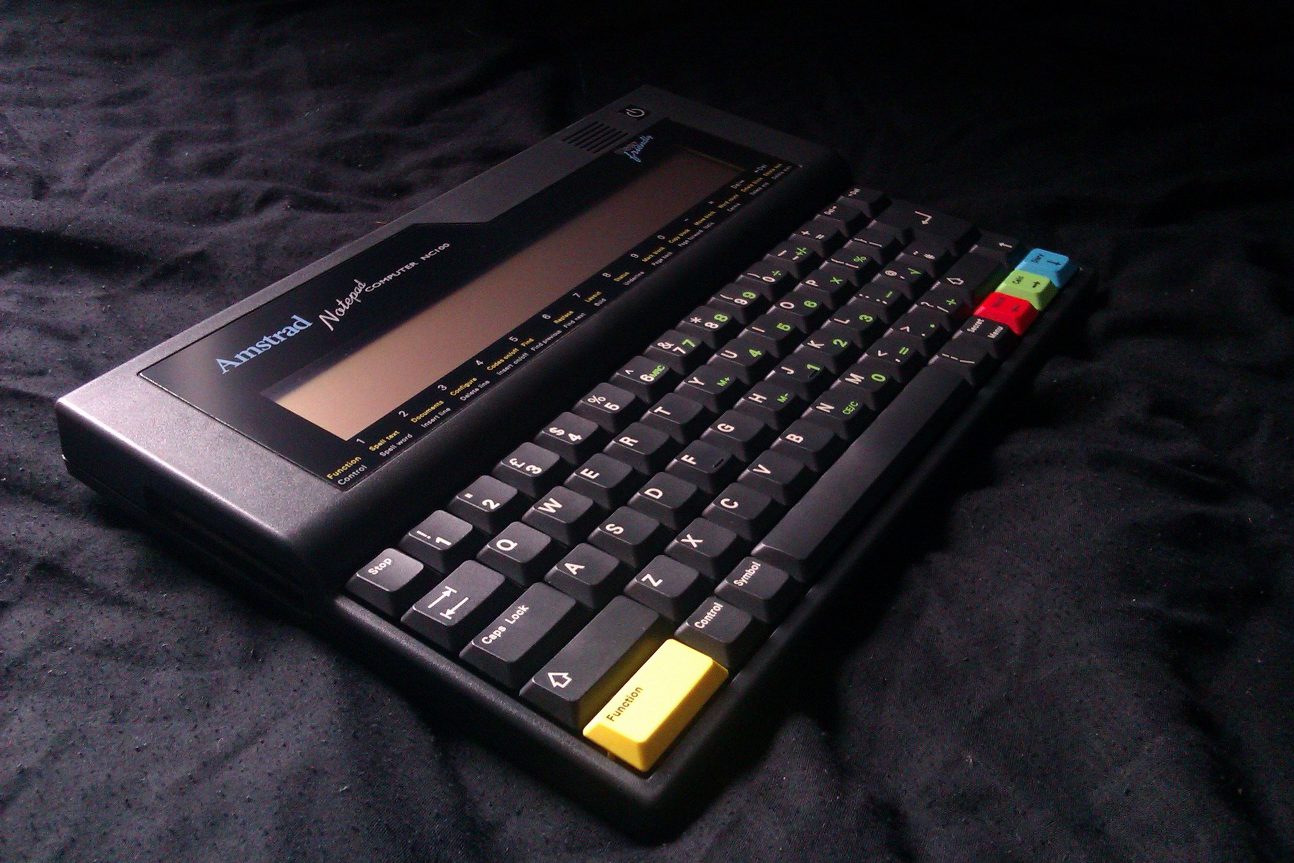 An Amstrad NC100 Notepad Computer, which is a flat device with an LCD screen and full keyboard. Most of the keys are black with white text, but four are brightly coloured: one is yellow, one is red, one is green, and one is blue.